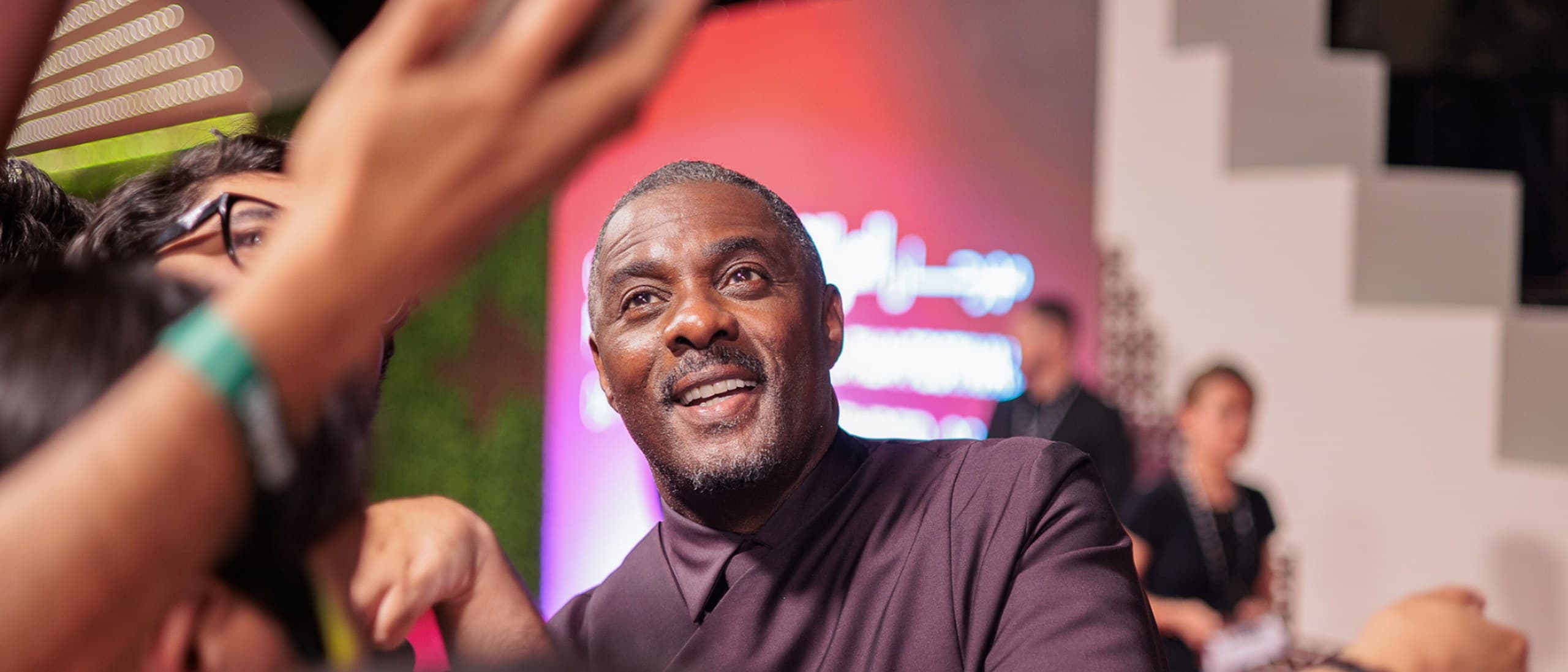 Idris Elba smiling as he greets fans holding up phones on the red carpet at the Red Sea International Film Festival 2025