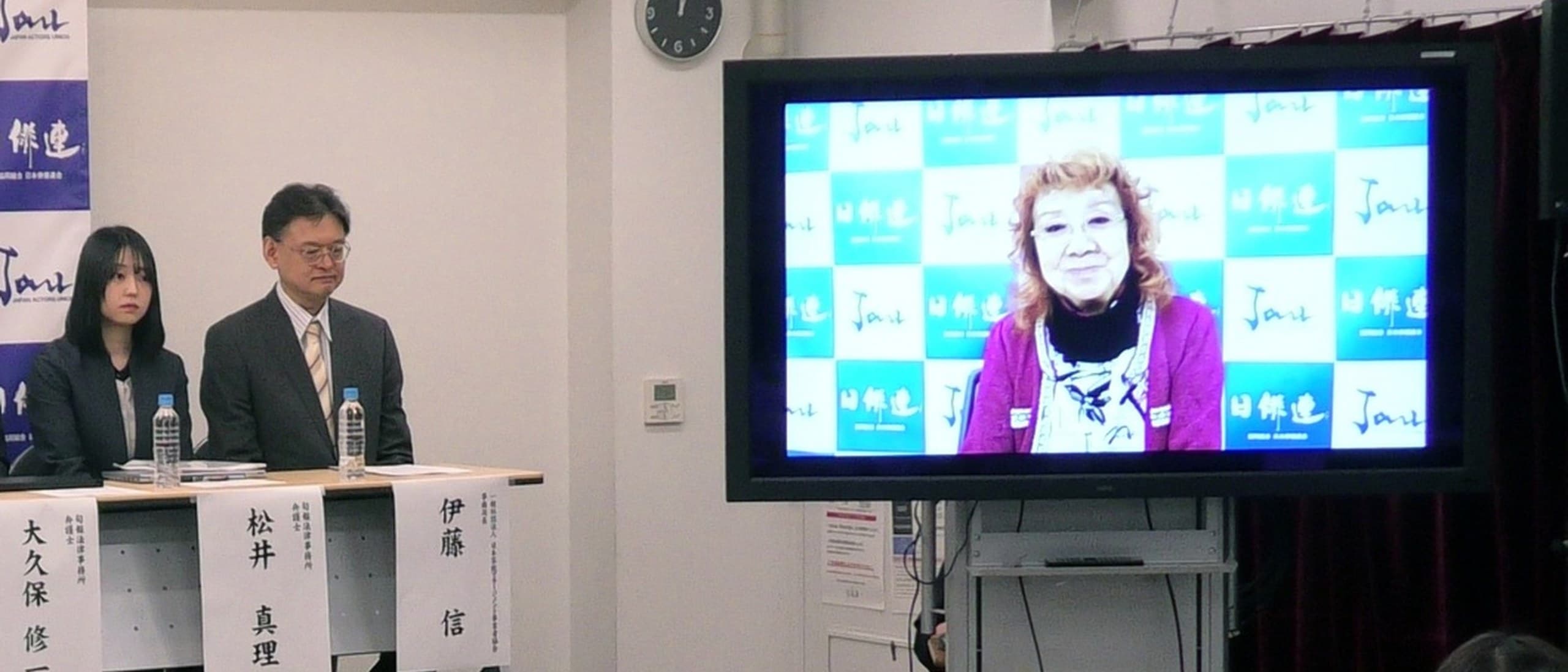 Masako Nozawa smiling on a large screen as she delivers a video message to the Japan Actors Union press conference launching its AI voice guidelines in Tokyo, with union officials seated at a table beside the screen