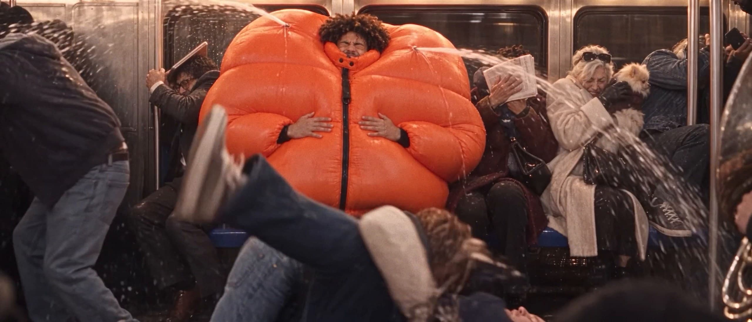 A man in a hugely inflated orange puffer jacket sits in a crowded New York subway car as jets of sweat spray across startled commuters, one falling in the foreground