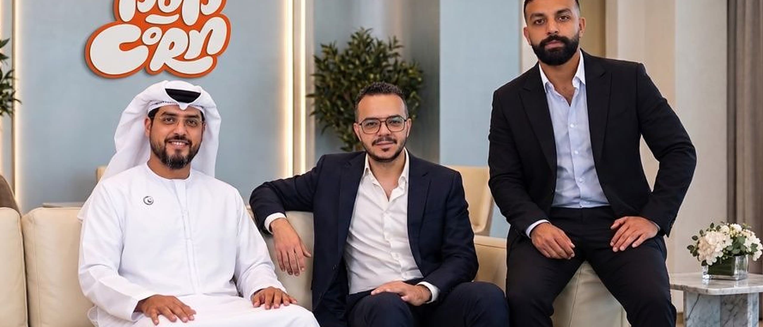Three men pose in front of an orange Popcorn logo on a pale blue wall, two seated on a cream sofa and one perched on its arm, one in a white kandura and ghutra and two in dark suits