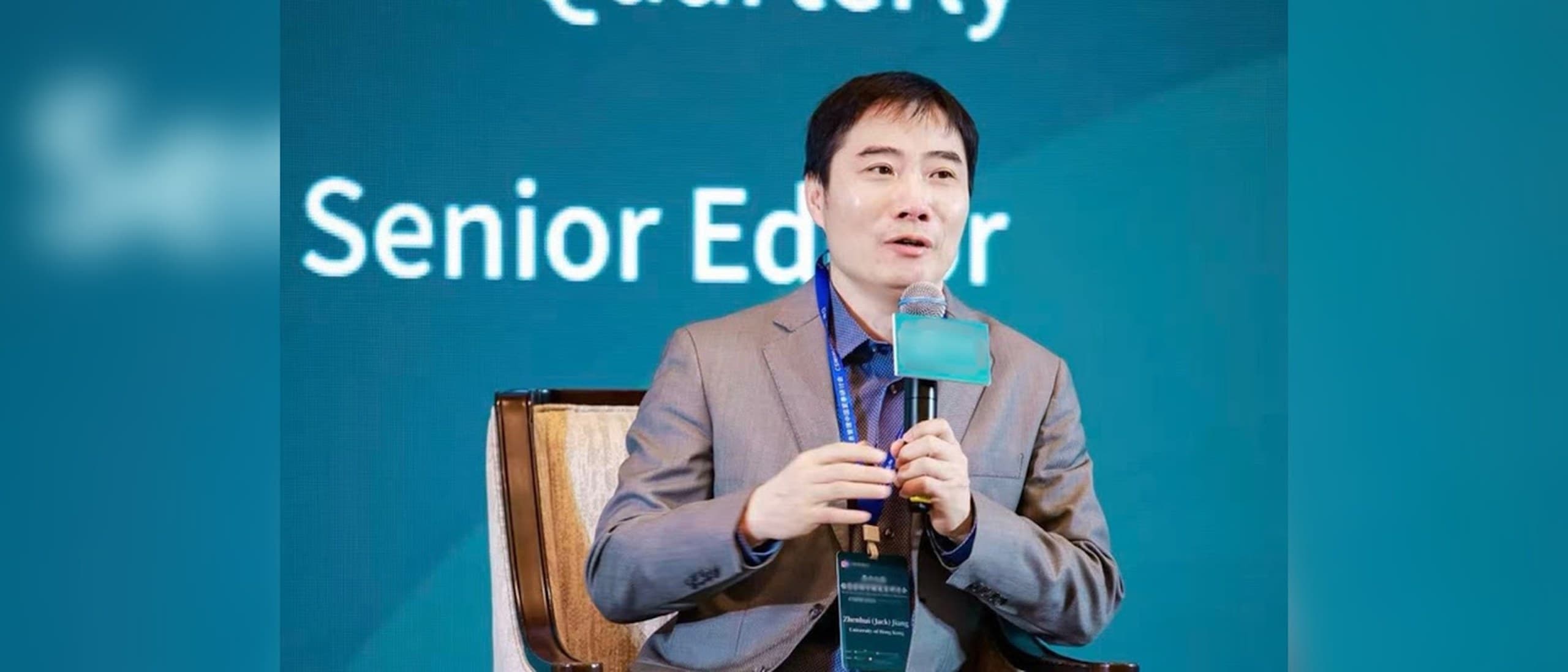 Professor Jack Jiang of HKU Business School, in a gray suit, speaks into a microphone while seated in an armchair on a conference stage