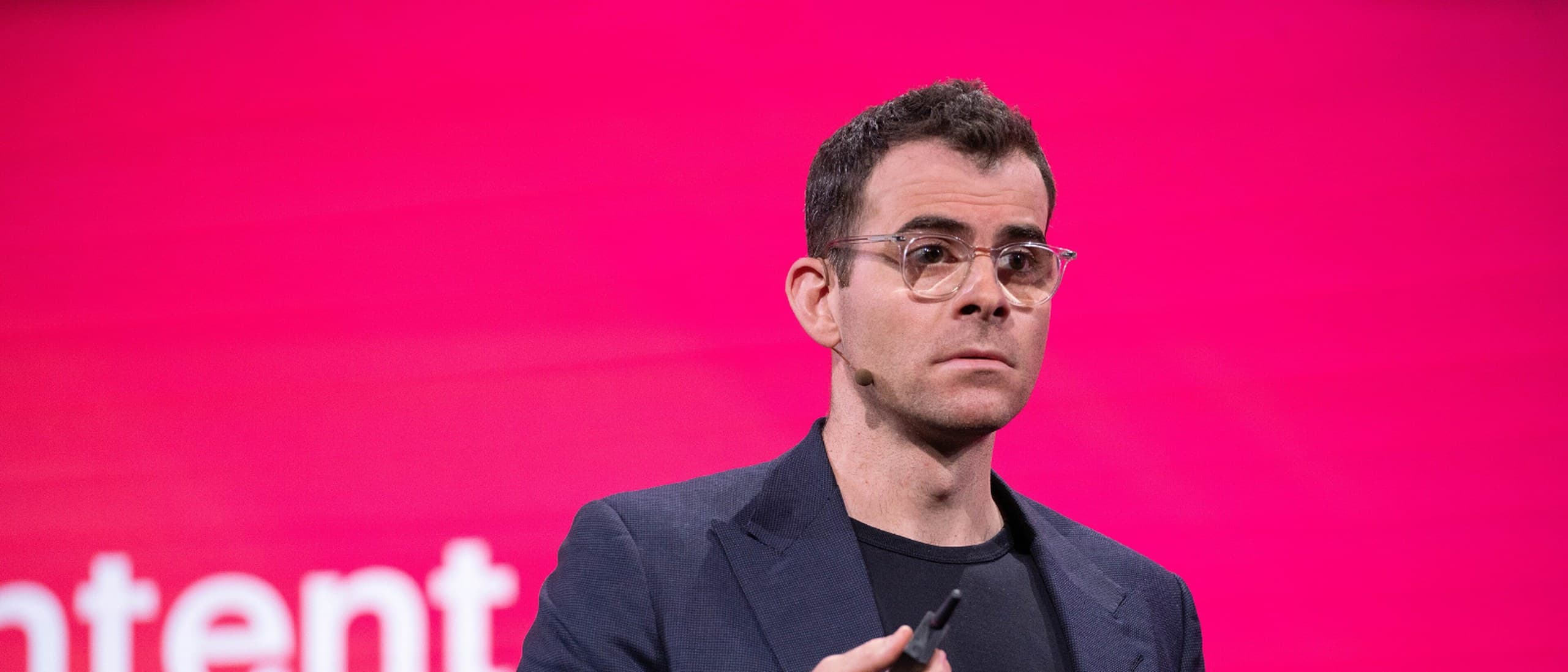 Adam Mosseri, head of Instagram, in glasses and a navy blazer, speaking on stage in front of a bright pink screen