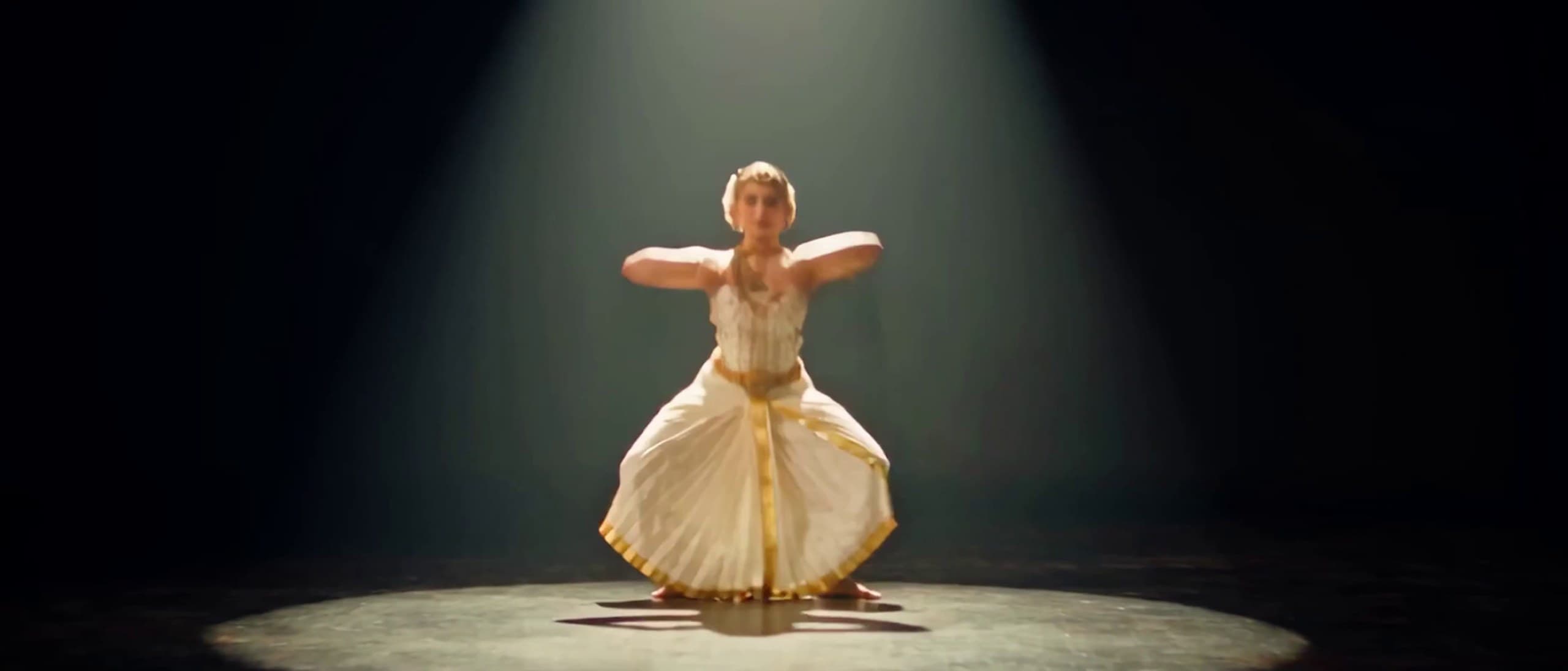 A dancer in a white and gold costume holds a deep half-seated pose, arms raised, inside the circle of a single spotlight on a black stage