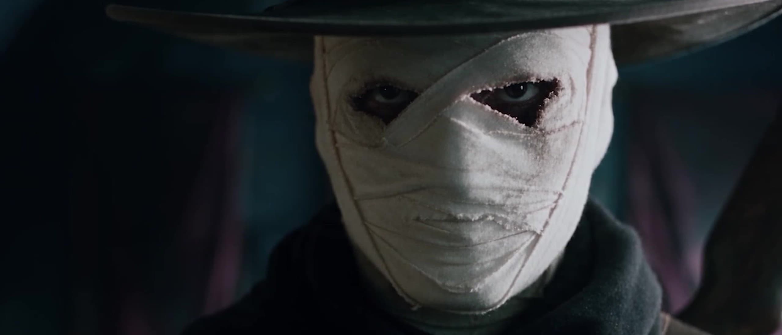 The Collector, a man with a bandaged white face under a wide-brimmed leather hat and dark scarf, stares into the camera in a dim hall