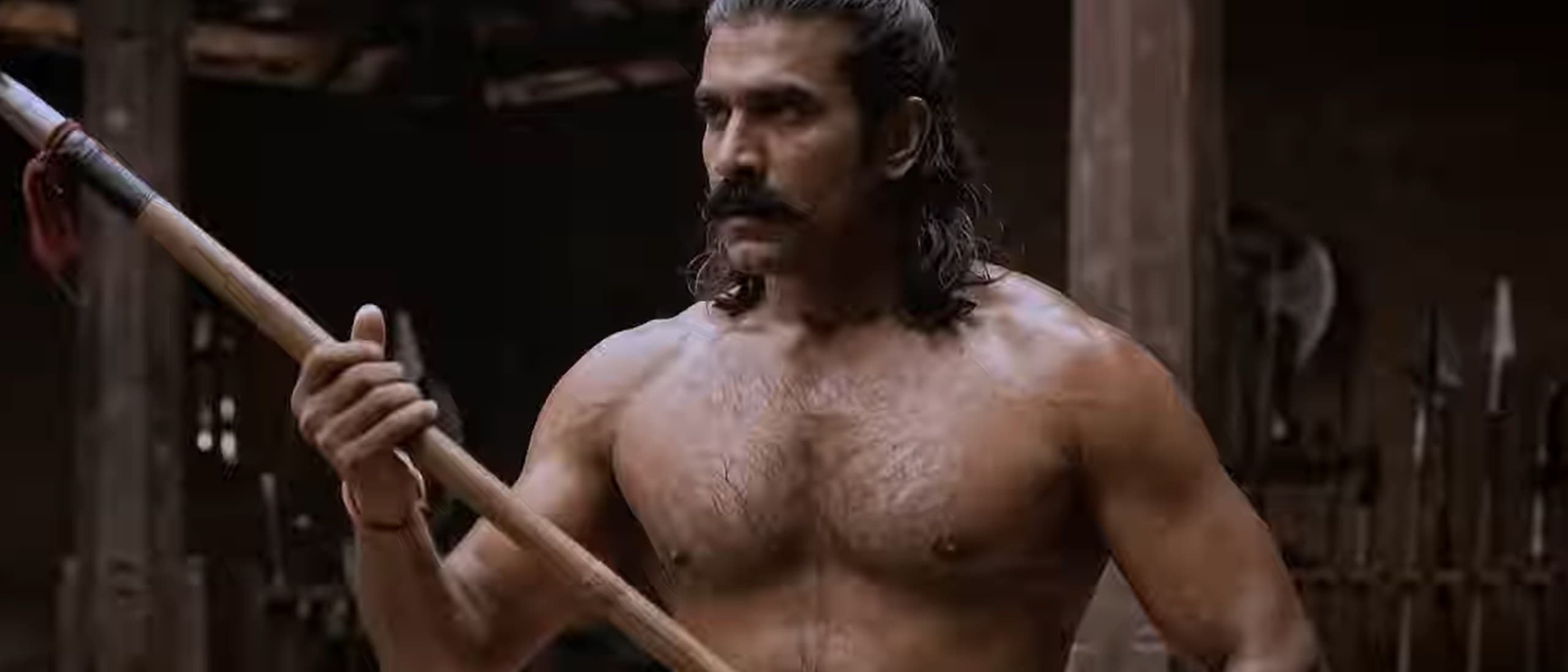 A still from the film Chekavar: a bare-chested warrior with a topknot and moustache grips a spear in a dim wooden armoury, with racks of blades behind him