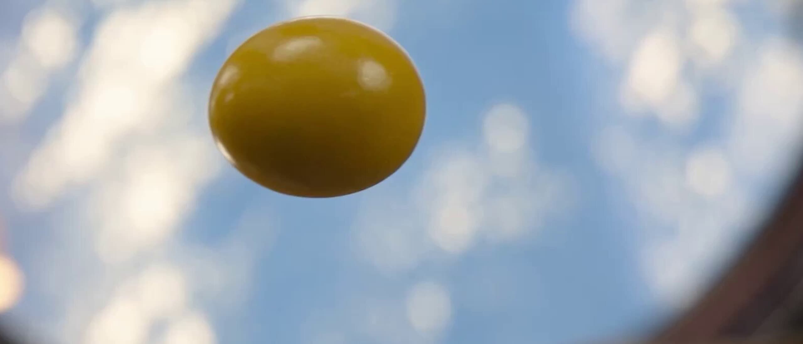 A yellow candy suspended in mid-air against a blue sky with blurred clouds, shot from below through a curved lens.