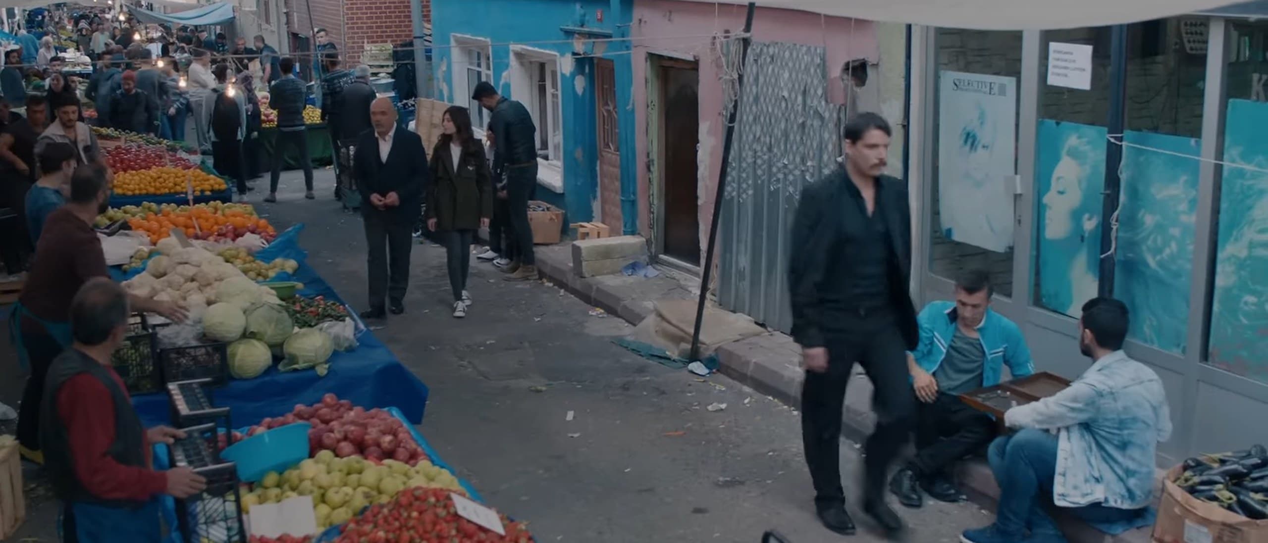 A busy street market in the Çukur neighbourhood of Istanbul, with produce stalls on the left and characters walking past painted houses, in a still from the original series