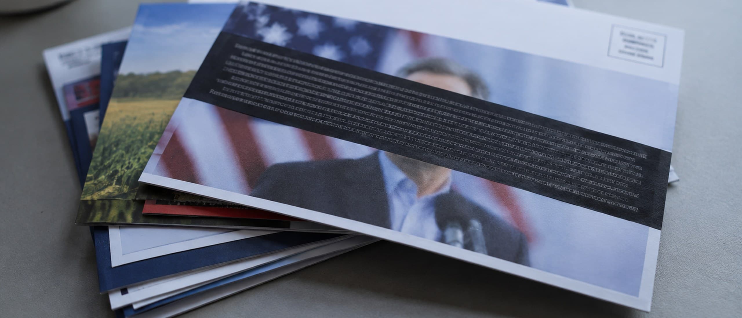 A stack of political campaign mailers on a grey desk, the top flyer showing a blurred candidate at a podium in front of a US flag with its text blacked out by a redaction bar