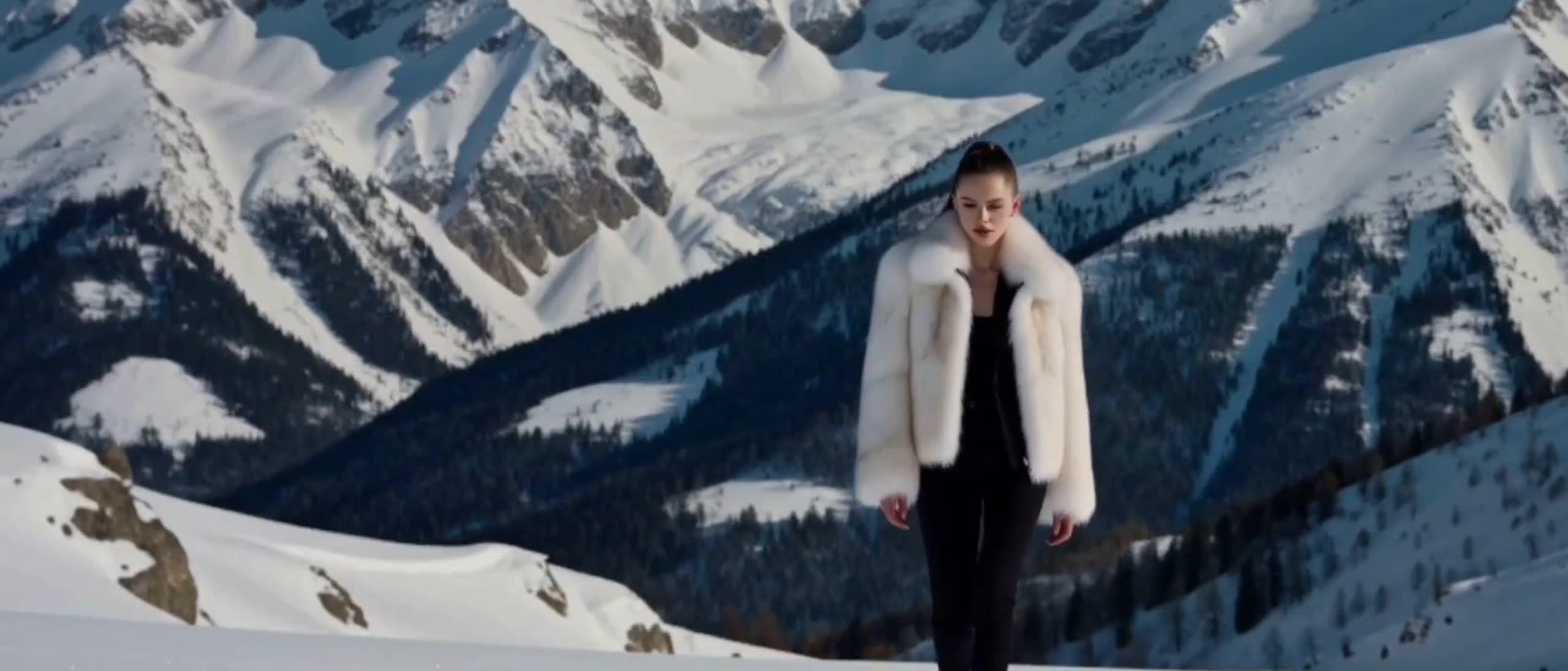 A woman in a white faux-fur jacket and black trousers walks toward the camera across fresh snow, with sunlit snowy mountain peaks and a dark pine valley behind her