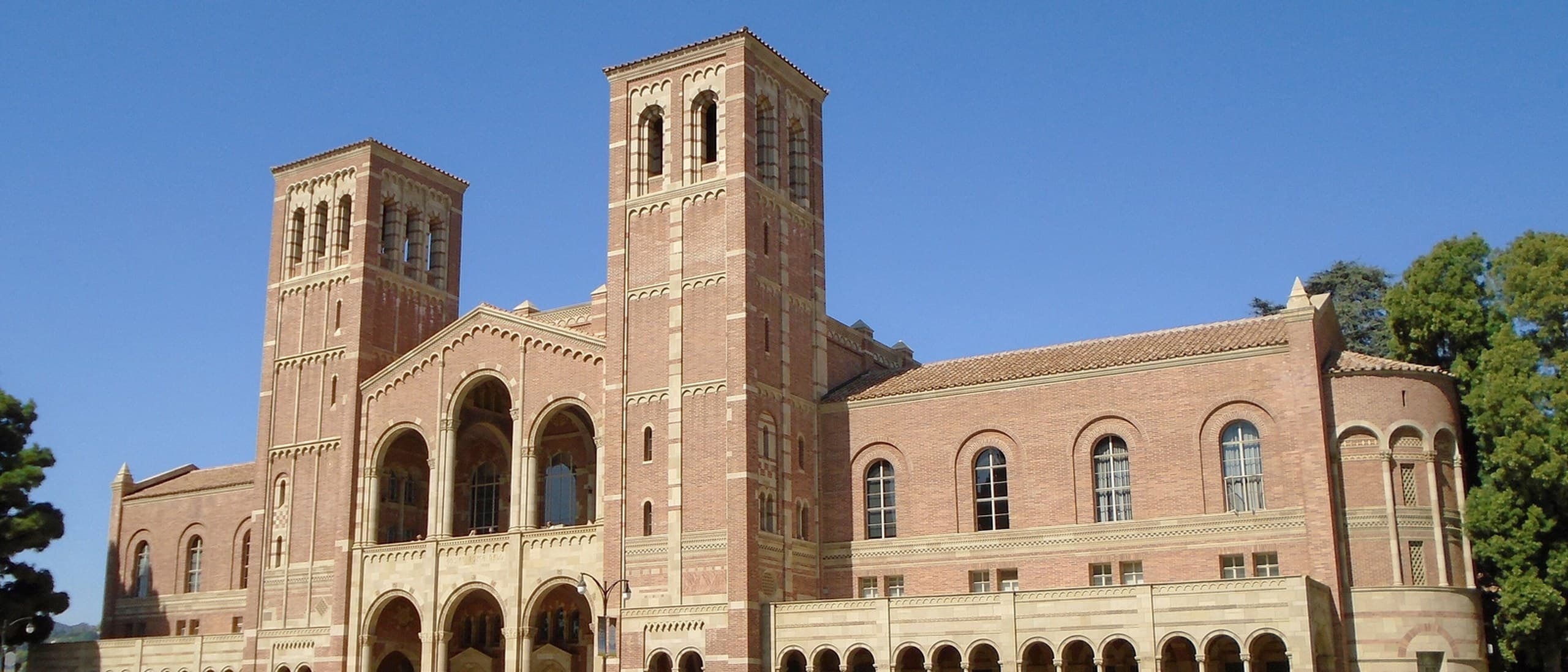 Royce Hall at UCLA, a red-brick building with two bell towers and arched loggias, under a clear blue sky above a lawn