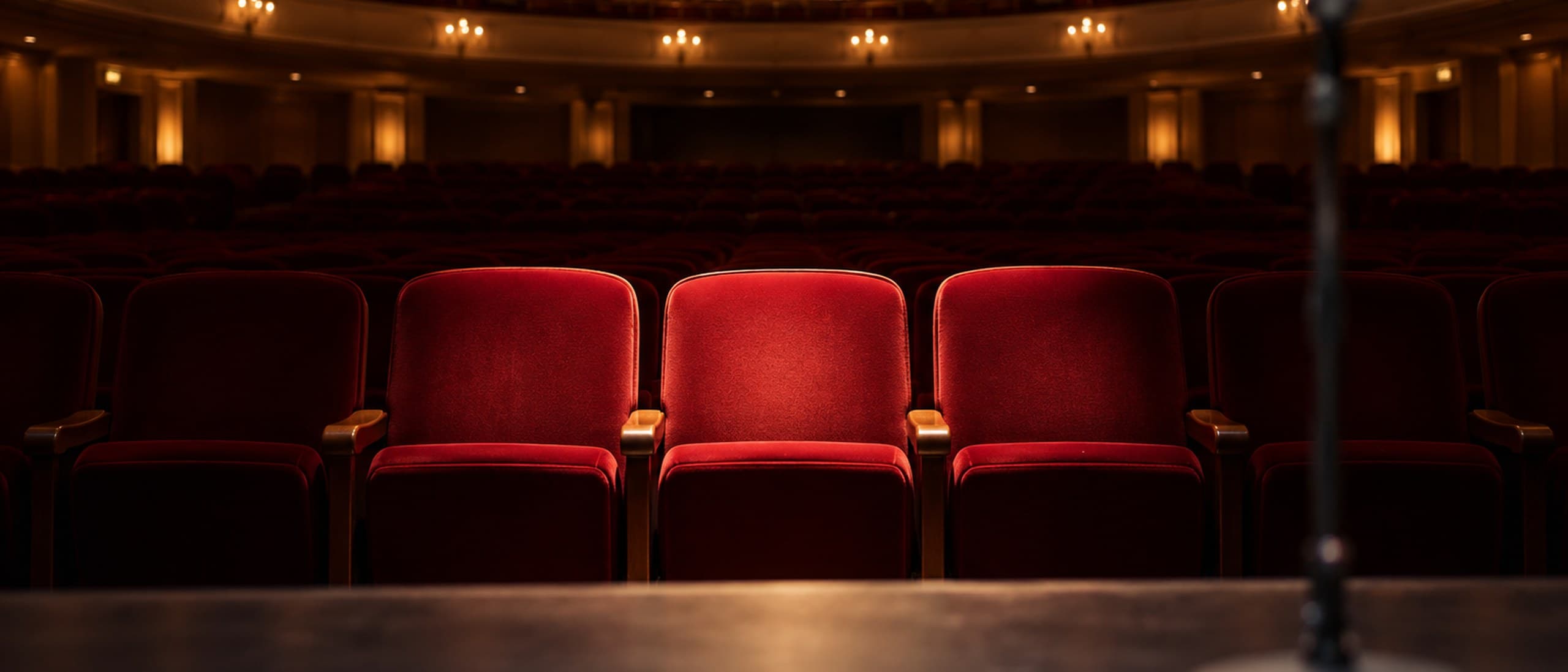 Three empty red velvet seats lit by a warm spotlight in the front row of a grand concert hall, with a microphone stand blurred in the foreground